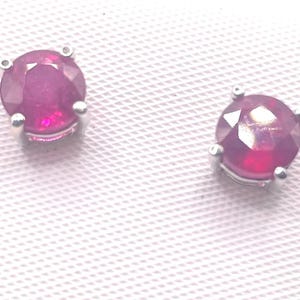 May include: Two round, ruby-colored gemstone earrings with silver settings. The gemstones have a faceted cut, reflecting light. The earrings are set against a white, textured background.