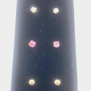 May include: Three pairs of stud earrings arranged vertically. The top pair features clear stones, the middle pair features round pink stones, and the bottom pair consists of plain gold-toned metal spheres. Each earring is secured with a prong setting.