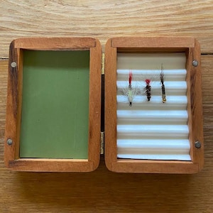 May include: Open wooden fly fishing box with a green interior on the left and a white corrugated interior on the right. The right side holds three colorful fishing flies. The box is open, revealing the contents.