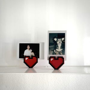 May include: Two Polaroid-style photo holders with red pixelated heart bases. One holds a photo of a couple, the other a dog. The holders are on a white shelf against a white wall.