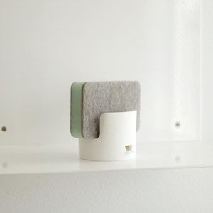 Peut inclure: Un support à éponge cylindrique blanc avec une ouverture incurvée, contenant une éponge rectangulaire grise et verte. Le support est sur une étagère blanche, créant une esthétique minimaliste. Le design est simple et fonctionnel.
