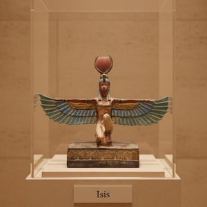 May include: A colorful statue of Isis, an ancient Egyptian goddess, displayed in a glass case. The statue features outstretched wings in blue and gold, a headdress, and is mounted on a rectangular base. The name "Isis" is displayed on a small plaque.
