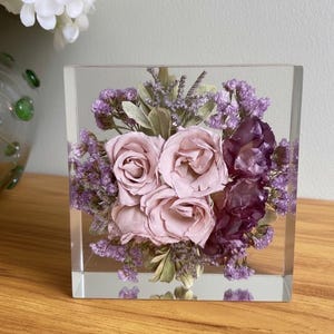 May include: A clear resin block, square in shape, contains a floral display. It features pale pink roses, deep purple flowers, and small purple blooms. Green foliage provides contrast. The block rests on a wooden surface.