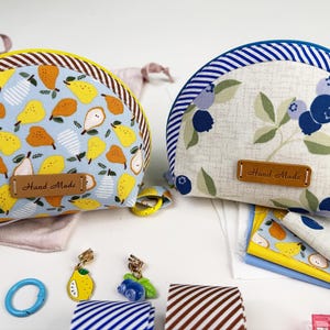 May include: Two handmade, half-circle pouches. One features a light blue background with yellow and brown pears. The other has a white background with blue blueberries and green leaves. Both have a brown leather tag that says "Hand Made".