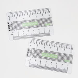 May include: Two clear rectangular rulers featuring black printed markings for both inches and centimeters. Each ruler displays a grid pattern in the center with the text MELBLOOM printed in white on a light green rectangular background.