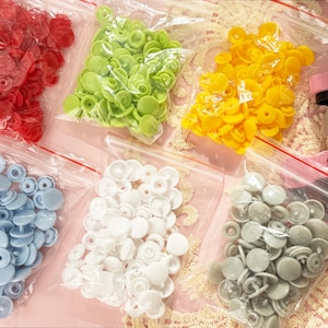 May include: A collection of colorful snap fasteners in clear plastic bags. The snaps are in various colors, including red, green, yellow, blue, white, and gray. A pink snap press tool is visible in the background, suggesting a sewing or crafting theme.