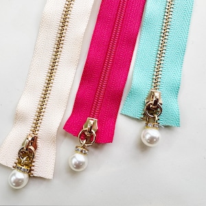 May include: Close-up of three zippers in cream, fuchsia, and teal. Each zipper has a gold-colored pull with a dangling pearl and rhinestone accent. The zippers are arranged diagonally against a white background.