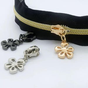 May include: A black zipper tape with gold-toned teeth and a gold-toned slider featuring a flower-shaped pull. Two additional loose zipper sliders are present, one in a matte black finish and one in a polished silver finish, both with flower-shaped pulls.