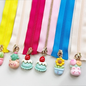 May include: A collection of colorful zippers in yellow, cream, pink, light pink, blue, and cream. Each zipper has a decorative charm, including a cactus, a bear with a cactus, two cakes with strawberries, a flower in a pot, and a bunny with flowers.