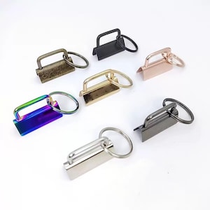 May include: A collection of seven metal fob hardware sets, each featuring a rectangular clamp and an attached circular split ring. The finishes include matte black, antique bronze, rose gold, polished gold, iridescent rainbow, gunmetal, and silver.