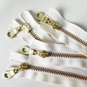 May include: Close-up of several ivory-colored zippers with brass-toned metal pulls and teeth. The zippers are arranged on a white surface, showcasing their design and construction. The YKK brand is visible on the pull.