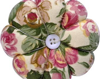 May include: A flower-shaped pin cushion with a white button center. The fabric petals feature a floral pattern in shades of pink, green, and cream. The petals are stitched together with white thread.