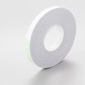 May include: A roll of white double-sided tape with a light green edge. The tape is on a white surface, and the roll is angled slightly, showing the inner core. The tape is likely used for crafts, scrapbooking, or general use.
