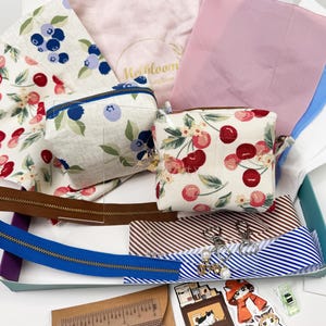 May include: A craft kit with fabric pieces, zippers, and small pouches. The fabric features cherry and blueberry patterns. Zippers are brown and blue. The kit includes a pink fabric with the text "Heirloom".
