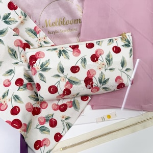 May include: A collection of sewing supplies featuring a white cherry-printed fabric, a matching zippered pouch, and a pink fabric. The pouch has a gold zipper. The text "Mellbloom Acrylic Templates" is visible.