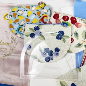 May include: An assortment of fabric pouches with fruit patterns, including pears, cherries, and blueberries. A clear acrylic template is in the foreground. A pink bag with the text "Melbloom Acrylic Templates" is also visible.