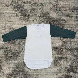 Könnte beinhalten: Weißes und dunkelgrünes Raglan-Baseball-Shirt mit langen Ärmeln. Das Shirt hat einen Henley-Ausschnitt mit drei Knöpfen. Das Shirt liegt flach auf einer strukturierten Oberfläche.