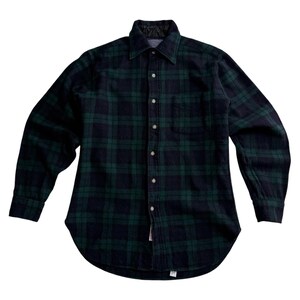 May include: A long-sleeved, button-down flannel shirt in a classic plaid pattern of dark green and navy blue. The shirt has a collared neckline and a curved hemline. The fabric appears to be a medium weight, suitable for cooler weather.