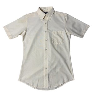 May include: A short-sleeved, button-down shirt in a light cream color. The shirt features a button-down collar, a single chest pocket, and a classic fit. The buttons are small and round, and the fabric appears to be a lightweight woven material.