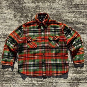 Pode incluir: Uma camisa de manga comprida com um padrão xadrez vermelho, verde, preto e branco. Possui dois bolsos no peito e uma gola preta. O tecido parece espesso e quente, ideal para temperaturas mais frias. A camisa é abotoada.