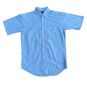 May include: A light blue short-sleeve button-down shirt. The shirt has a button-down collar, a chest pocket, and white buttons. The shirt is made of a lightweight fabric and is spread out on a white surface.