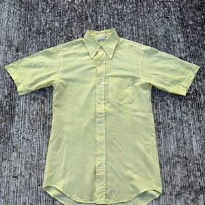 Puede incluir: Una camisa amarilla pálida de manga corta con botones y un bolsillo en el pecho. La camisa tiene un cuello clásico y está hecha de una tela ligera. La camisa está tendida sobre una superficie texturizada.