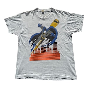 Può includere: T-shirt grigio chiaro con una grafica di Batman in volo, con un segnale del pipistrello giallo e nero e uno skyline arancione e nero. Il supereroe è raffigurato con un mantello blu e un costume grigio.