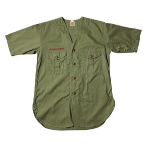 May include: Olive green short-sleeved shirt with a V-neck and button-down front. Features two chest pockets and the text "BOY SCOUTS - AMERICA" in red. The shirt has a curved hem and a small tag at the neckline.