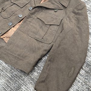Puede incluir: Chaqueta militar verde oliva vintage con cuatro bolsillos delanteros y botones. La chaqueta tiene un cuello con solapas y un forro interior marr&oacute;n. La tela muestra signos de desgaste, lo que se suma a su est&eacute;tica antigua.