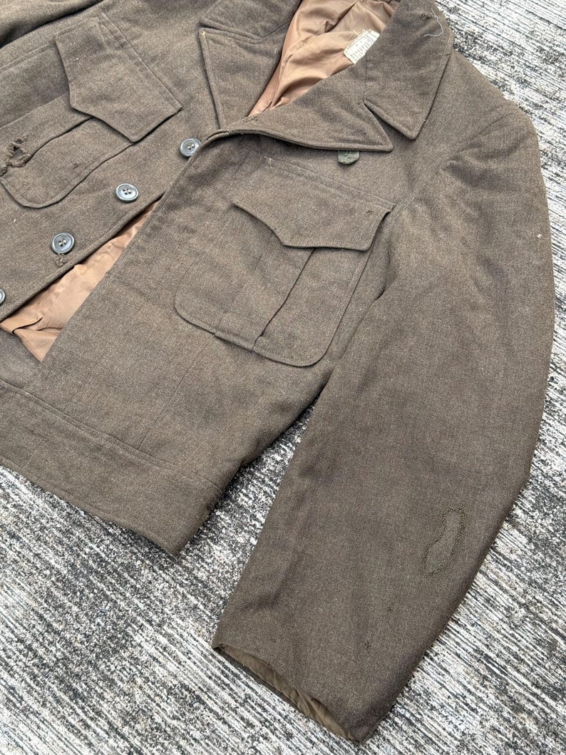 Puede incluir: Chaqueta militar verde oliva vintage con cuatro bolsillos delanteros y botones. La chaqueta tiene un cuello con solapas y un forro interior marr&oacute;n. La tela muestra signos de desgaste, lo que se suma a su est&eacute;tica antigua.