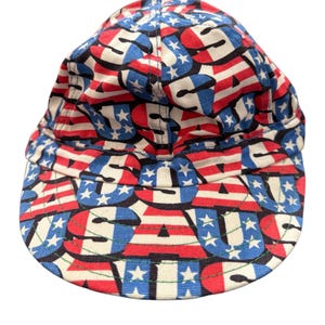 Könnte beinhalten: Patriotische Baseballkappe mit einem sich wiederholenden Muster aus roten und weißen Streifen, blauen Sternen und den Buchstaben "USA". Die Kappe hat einen gebogenen Schirm und eine runde Krone.