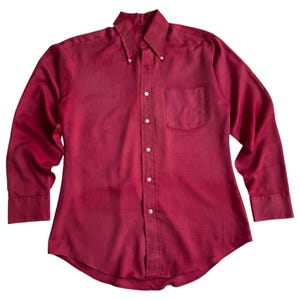 May include: A long-sleeved button-down shirt in a deep burgundy red color. The shirt features a pointed collar, a single chest pocket on the left side, and a full-length front button placket with white buttons. The fabric has a visible texture.