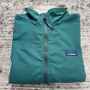 Puede incluir: Chaqueta L.L.Bean de color verde azulado con cremallera completa y cuello gris claro. La chaqueta tiene un pequeño parche rectangular con el logotipo de L.L.Bean y un tirador de cremallera con el nombre de la marca. La chaqueta está doblada.