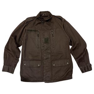 May include: A dark brown field jacket featuring a high collar, a central button-down placket, and long sleeves. The front includes two vertical zippered chest pockets, two large lower flap pockets with buttons, and a small rectangular patch.
