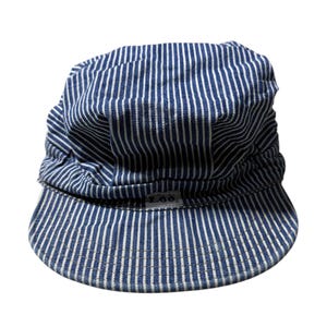 Puede incluir: Gorra de ferrocarril a rayas azules y blancas. La gorra tiene una corona redondeada y una visera corta y curvada. La tela es un material similar al denim con rayas verticales. El logotipo de Lee es visible en la visera.