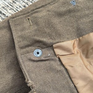 Puede incluir: Primer plano de una chaqueta marr&oacute;n con un bot&oacute;n que dice "UNITED CARR". La chaqueta tiene un forro marr&oacute;n claro y un peque&ntilde;o dise&ntilde;o bordado en azul claro. La tela tiene un aspecto texturizado.