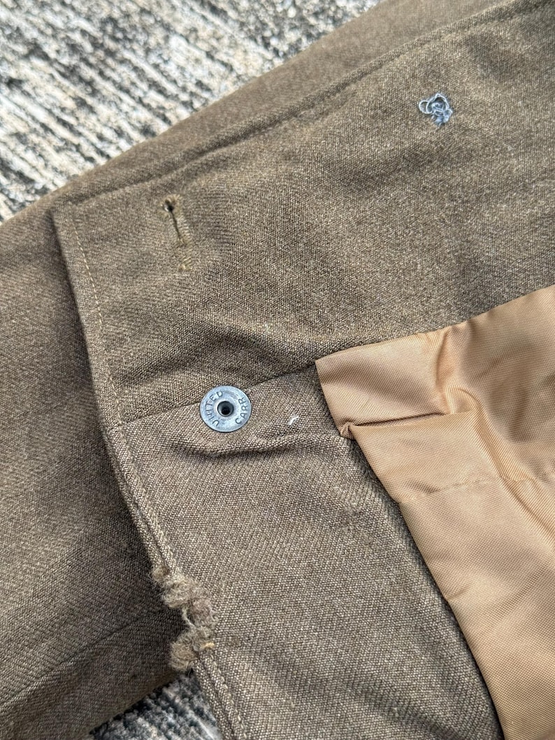 Puede incluir: Primer plano de una chaqueta marr&oacute;n con un bot&oacute;n que dice "UNITED CARR". La chaqueta tiene un forro marr&oacute;n claro y un peque&ntilde;o dise&ntilde;o bordado en azul claro. La tela tiene un aspecto texturizado.