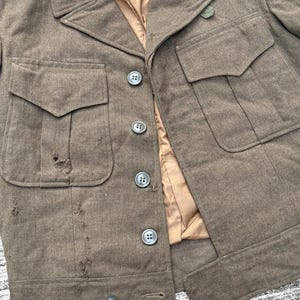 Puede incluir: Chaqueta militar verde oliva vintage con cuatro bolsillos delanteros y botones. La chaqueta tiene un cuello con solapas y un forro beige. La tela muestra signos de desgaste, con algunos agujeros y deshilachados.