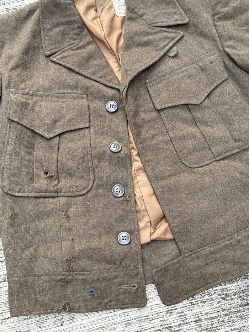 Puede incluir: Chaqueta militar verde oliva vintage con cuatro bolsillos delanteros y botones. La chaqueta tiene un cuello con solapas y un forro beige. La tela muestra signos de desgaste, con algunos agujeros y deshilachados.