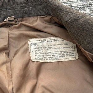 Puede incluir: Primer plano del interior de una chaqueta de lana verde oliva vintage. El forro es de color beige claro. Una etiqueta con texto est&aacute; cosida dentro de la chaqueta, proporcionando instrucciones de ajuste. El cuello de la chaqueta es visible en la parte superior.