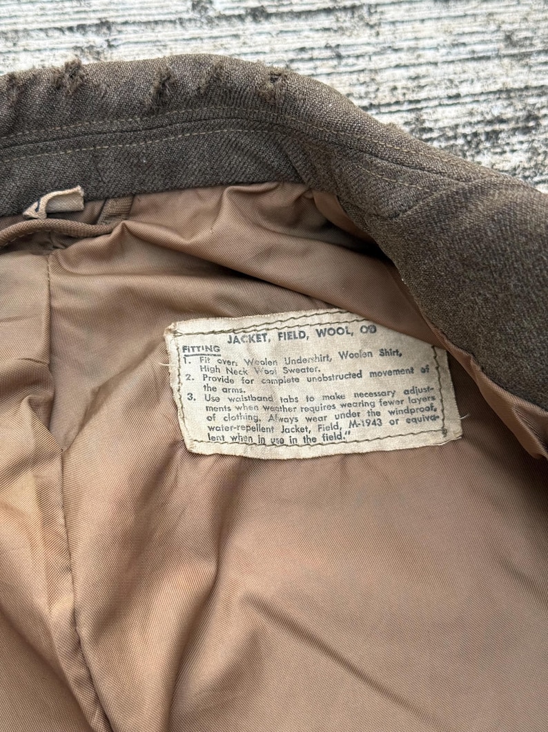 Puede incluir: Primer plano del interior de una chaqueta de lana verde oliva vintage. El forro es de color beige claro. Una etiqueta con texto est&aacute; cosida dentro de la chaqueta, proporcionando instrucciones de ajuste. El cuello de la chaqueta es visible en la parte superior.