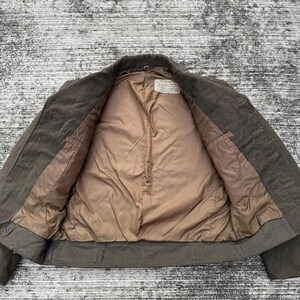 Puede incluir: Chaqueta verde oliva abierta, con forro interior beige. La chaqueta tiene cierre de botones y dos bolsillos delanteros. El forro interior es de tela lisa beige. La chaqueta parece ser de lana o mezcla de lana.