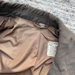 Puede incluir: Primer plano del interior de una chaqueta verde oliva vintage. El forro es de color marr&oacute;n claro. La etiqueta dice "34R" y una etiqueta m&aacute;s peque&ntilde;a con texto sobre el ajuste de la chaqueta. La chaqueta parece ser de lana.