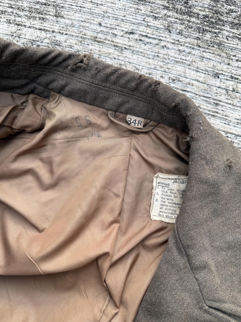 Puede incluir: Primer plano del interior de una chaqueta verde oliva vintage. El forro es de color marr&oacute;n claro. La etiqueta dice "34R" y una etiqueta m&aacute;s peque&ntilde;a con texto sobre el ajuste de la chaqueta. La chaqueta parece ser de lana.