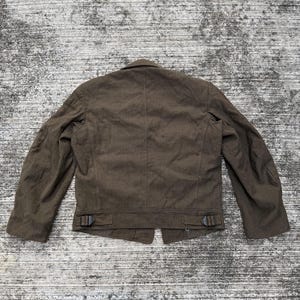 Puede incluir: Chaqueta marr&oacute;n con cierre de botones y mangas largas. La chaqueta tiene dos peque&ntilde;as hebillas en la parte inferior de la espalda y un cuello con solapas. La tela parece ser un tejido texturizado.