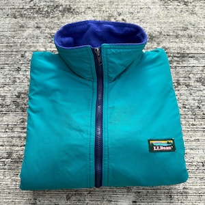 Puede incluir: Chaqueta reversible azul verdoso y azul con cierre de cremallera completo. La chaqueta tiene un forro polar azul y un exterior azul verdoso. El cuello es de estilo alzado. El logotipo de LLBean está en la parte inferior izquierda de la chaqueta.