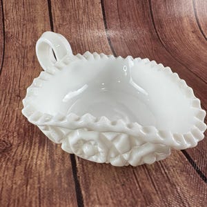 May include: A white, oval-shaped dish with a handle. The dish has a ruffled edge and a decorative floral pattern on the base. The dish is made of a glossy, opaque material.