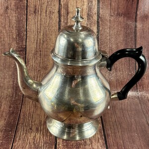 May include: A silver teapot with a black handle and a decorative finial on the lid. The teapot has a bulbous body and a curved spout. The background is a wooden surface.