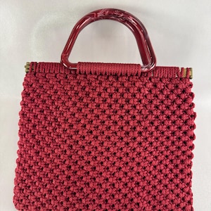 May include: A red macrame handbag with a rectangular shape. The bag features a woven texture and a curved, marbled red handle. The bag's frame is a brass color.
