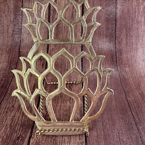 May include: A silver-colored metal decorative plate holder with an openwork floral design. The holder is made of two connected sections, with a chain connecting the top and bottom. The holder is on a wooden surface.